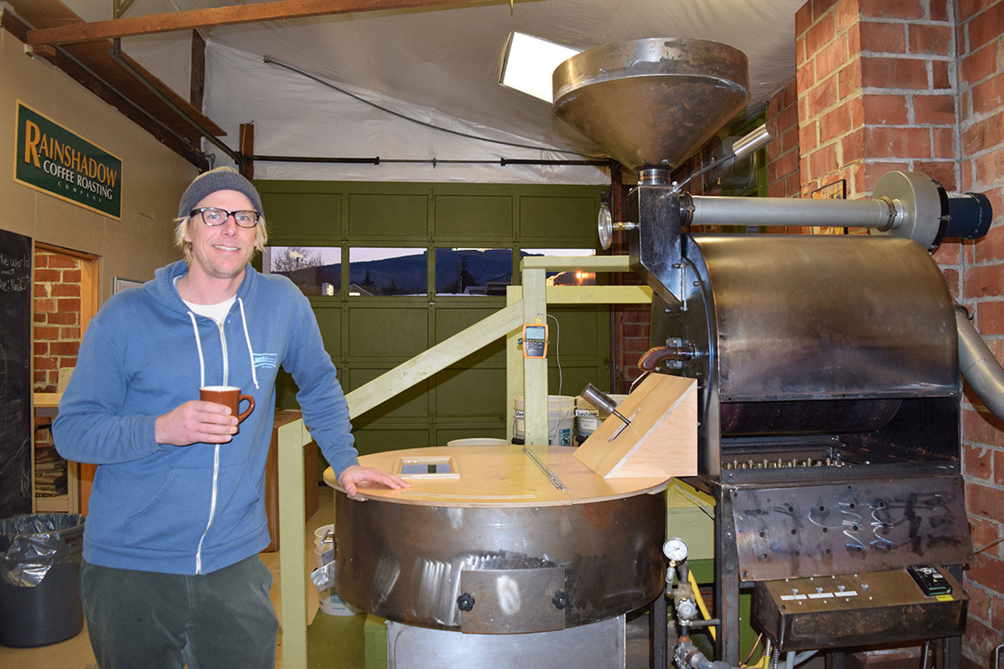 Nick Batcheller of Rainshadow Coffee Roasting Co. stands with the roasting machine his father and brother built when the business first bought the space at 157 W. Cedar St. in Sequim. The family continues to roast and distribute coffee out of Sequim and plans to open a new roasting facility in Paso Robles, Calif. (Erin Hawkins/Olympic Peninsula News Group)
