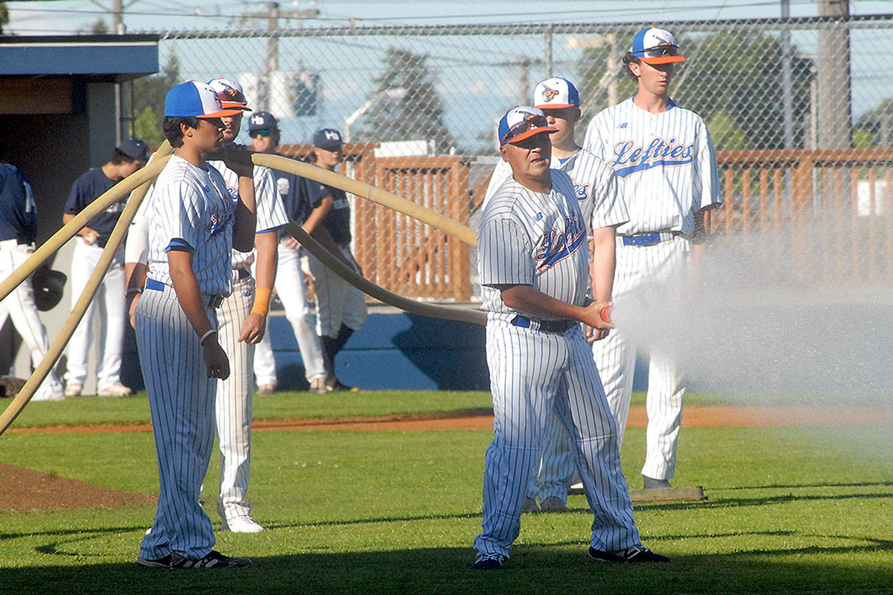 Members of the Port Angeles Lefties baseball team, including assistant coach Anthony Murillo, holding a hose, water down the infield prior to the start of Tuesday evening’s season opener against the Highline Bears at Port Angeles Civic Field. (Keith Thorpe/Peninsula Daily News)