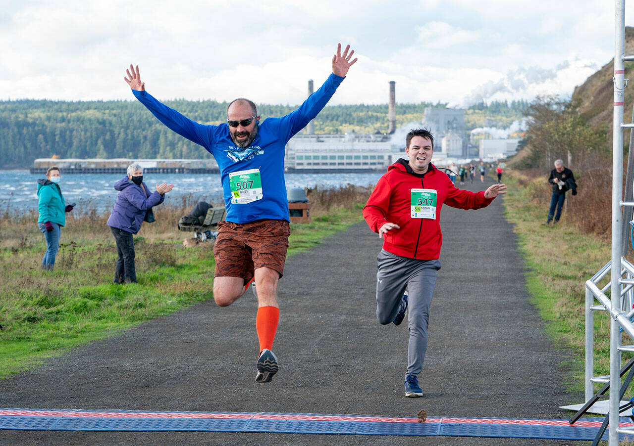 Sequim’s Russ Britton (507) jumps through the finish line after he completes the Run the Peninsula 5K on the Larry Scott Trail in Port Townsend on Saturday. Taran Johnson, (547) also from Sequim, races to the finish. (Steve Mullensky/for Peninsula Daily News)