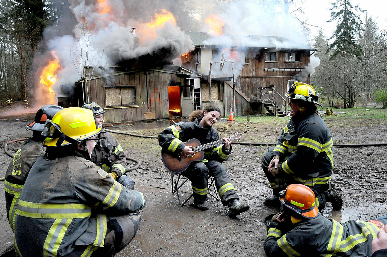 Clallam County Fire District 2 firefighter Mya Delano plays her guitar surrounded by fellow squad members as a house at 141 Harry Brown Road burns behind her on Sunday west of Port Angeles. The house was intentionally torched as a training exercise, giving firefighters an opportunity to practice in a live fire situation. Fire districts 2 and 4, along with members of the Port Angeles Fire Department, took part in the event. (Keith Thorpe/Peninsula Daily News)