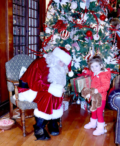Santa, played by Alexis Smith, a Lake Crescent Lodge employee, wonders why Zoey Doud, 3, of Port Angeles thinks the big present under the tree is for her. Santa turned on the outdoor lights at the lodge on Saturday then sat down by the fireplace to visit with the many children who came to the festivities. Entertainment was provided by the holiday songs of the Grand Olympic Chorus. (Dave Logan/for Peninsula Daily News)