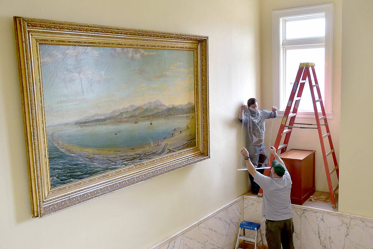 David Brownell, executive director of the North Olympic History Center, top, takes a piece of ultraviolet-filtering window tinting from Ralph Parsons, Clallam County maintenance worker, in an effort on Tuesday to protect historic paintings on the stairway of the section of the county courthouse, including an 1890s depiction of Port Angeles Harbor by artist John Gustaf Kalling. The history center is working with the county to preserve the stairway artworks by adding the window coatings to reduce damage from sunlight and installing an electronic UV monitor to track potentially harmful rays. (Keith Thorpe/Peninsula Daily News)