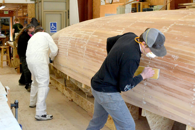 From left to right, Northwest School of Wooden Boatbuilding students Krystol Pasecznyk and Scott McNair sand a Prothero Sloop with Sean Koomen, the school’s boat building program director. Koomen said the sanding would take one person a few days. He said the plan is to have 12 people sand it together, which will take a few hours. (Elijah Sussman/Peninsula Daily News)