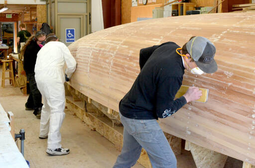 From left to right, Northwest School of Wooden Boatbuilding students Krystol Pasecznyk and Scott McNair sand a Prothero Sloop with Sean Koomen, the school’s boat building program director. Koomen said the sanding would take one person a few days. He said the plan is to have 12 people sand it together, which will take a few hours. (Elijah Sussman/Peninsula Daily News)
