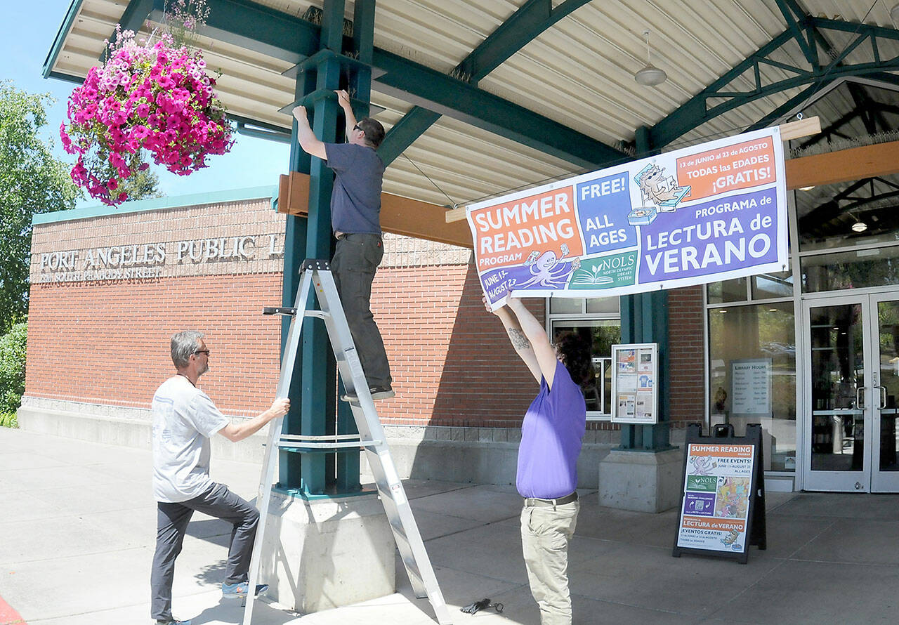 Maintenance workers, from left, Brian Phillips, Jeff Clark and Noah Mohmand, suspend a banner outside the Port Angeles Public Library to gather interest in the library system’s Summer Reading Program, which runs from Friday through Aug. 23. The program offers free books and prizes for avid readers at the system’s branches in Port Angeles, Sequim, Forks and Clallam Bay. (Keith Thorpe/Peninsula Daily News)