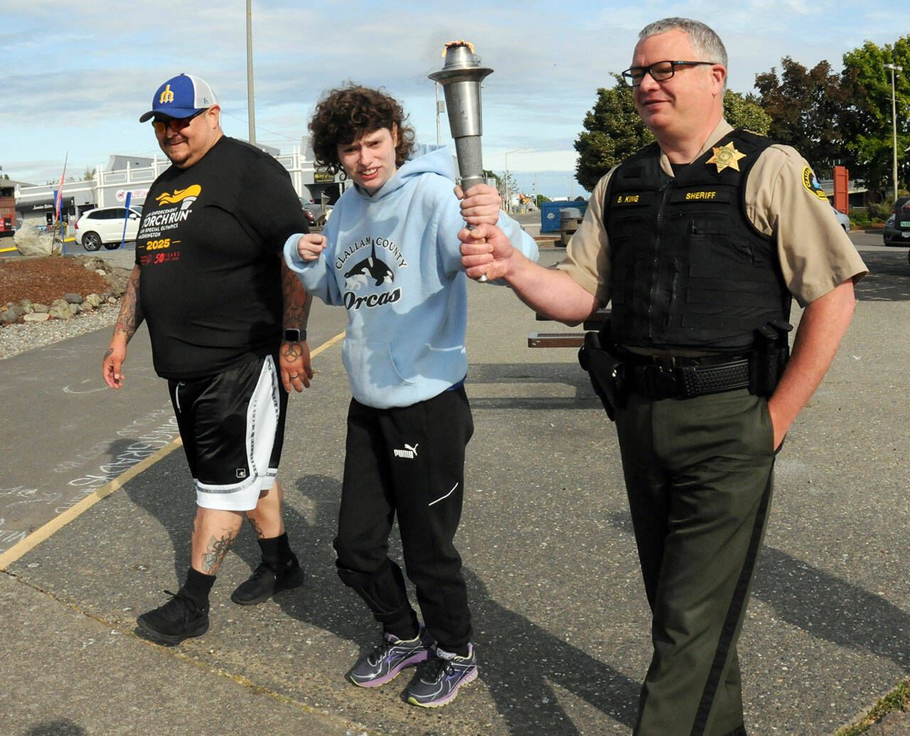 Special Olympian Deni Isett, center, holds a ceremonial torch with Clallam County Sheriff Brian King, right, accompanied by Lt. Jim Thompson of the Lower Elwha Klallam Tribal Police on a leg of the Law Enforcement Torch Run on the Olympic Discovery Trail at Port Angeles City Pier. Tuesday’s segment of the run, conducted mostly by area law enforcement agencies, was organized to support Special Olympics Washington and was to culminate with a community celebration at 7 Cedars Casino in Blyn. (Keith Thorpe/Peninsula Daily News)